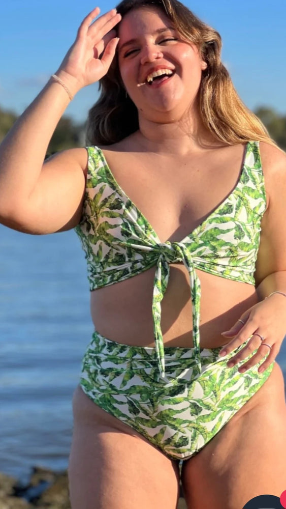 Model is smiling and wearing a mid coverage bikini bottom and a tie at the front bikini top. The pattern is made of green leas on a white base. The female model is on the beach. 