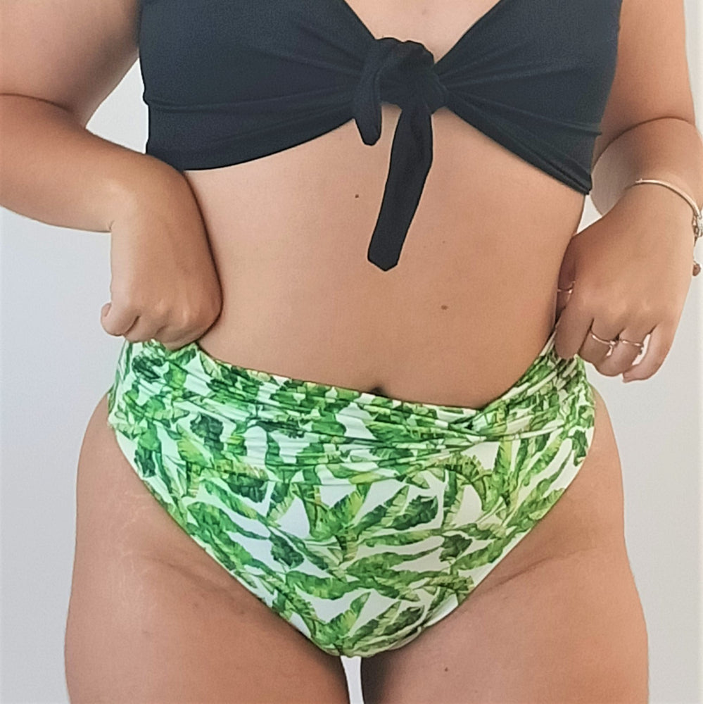 curvy model is wearing a high waisted bikini bottom with white background and with leaves in the pattern. black top